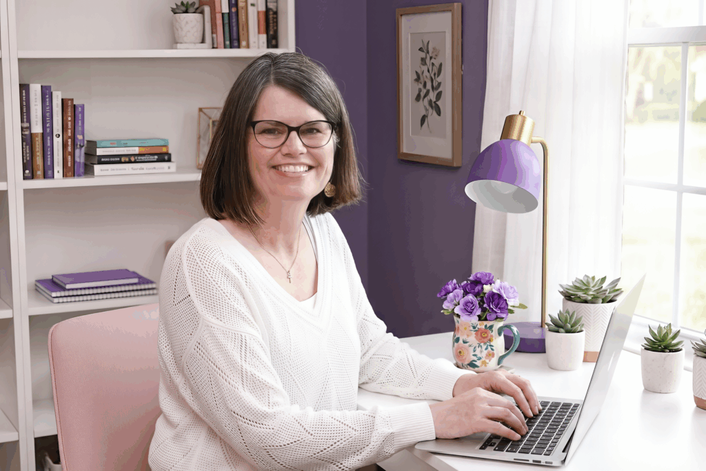 Gjoa Crandell, Founder of AI Training HQ, smiling and working at her laptop in a home office with purple accents.